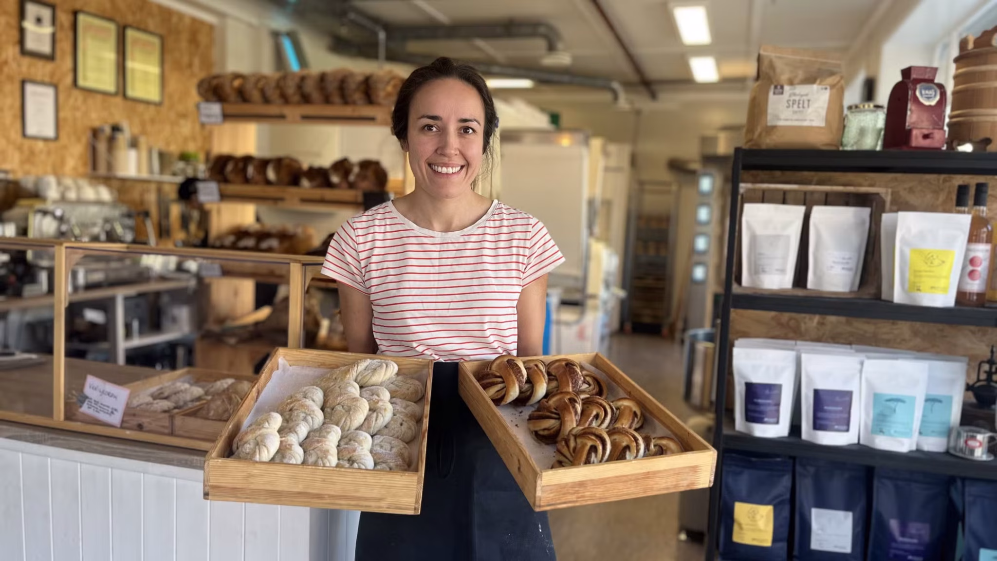 Smiling bakery worker presents two wooden trays of assorted bread and pretzel pastries in a bakery shop.