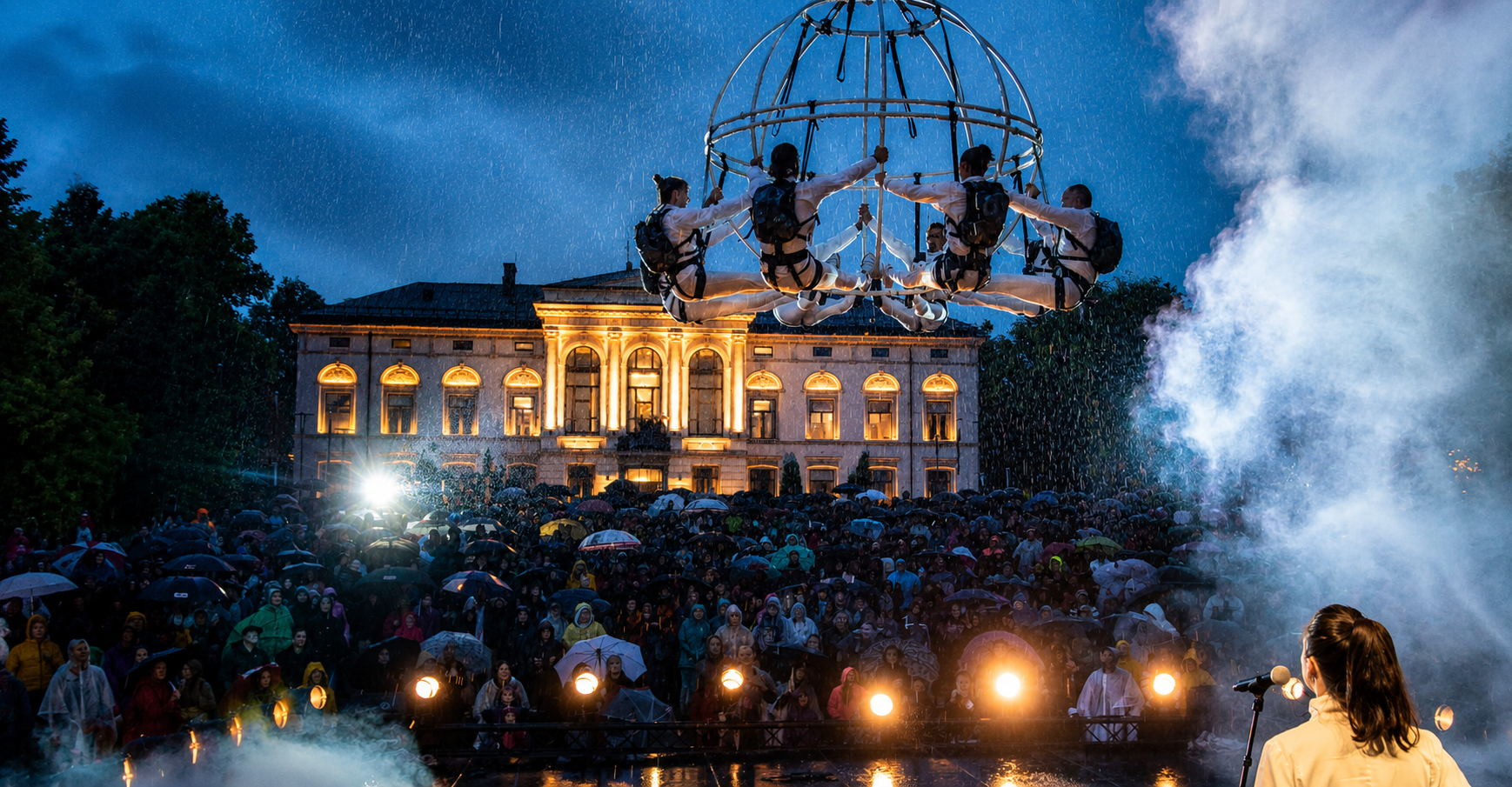 Aerial performance: four climbers hang inside a glowing circular rig above a crowded outdoor stage in the rain at night.