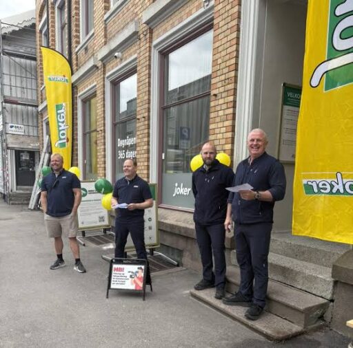 Four men in dark polo shirts stand outside a brick storefront with yellow banners and balloons, handing out papers at a storefront event.